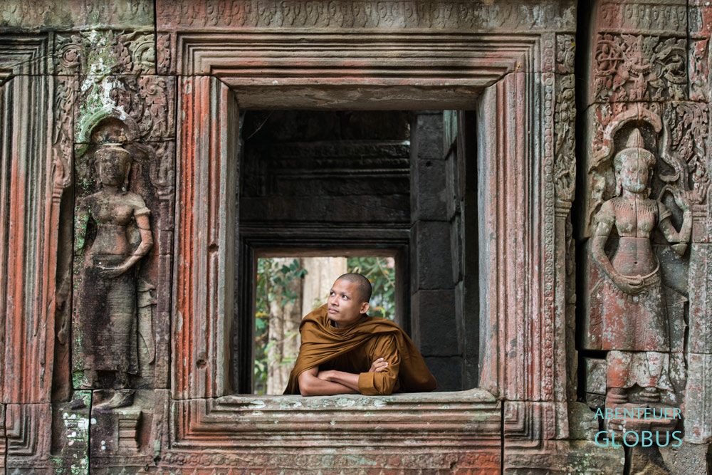 Angkor: Ein Mönch schaut in der Tempelruine Banteay Kdei aus dem Fenster, das von zwei Apsara-Tänzerinnen flankiert wird.  