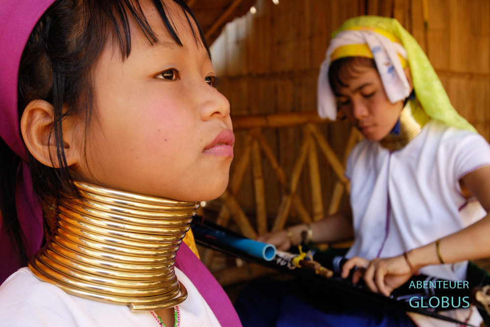 Mädchen der Padaung mit Messingspirale, im Hintergrund eine Frau beim Weben.