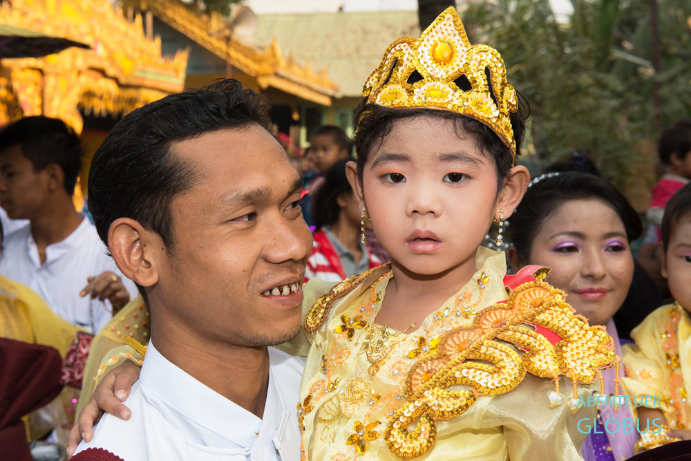 Zur Novizenweihe Shin Pyu nahe Mandalay trägt ein stolzer Vater seine Tochter in einem Seidenkleid auf dem Arm.