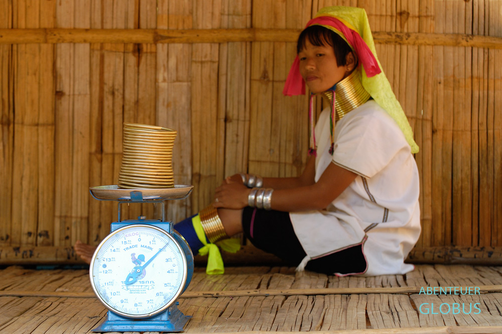 Waage mit Messing-Halsspirale im Vordergrund, im Hintergrund eine sitzende Padaung-Frau