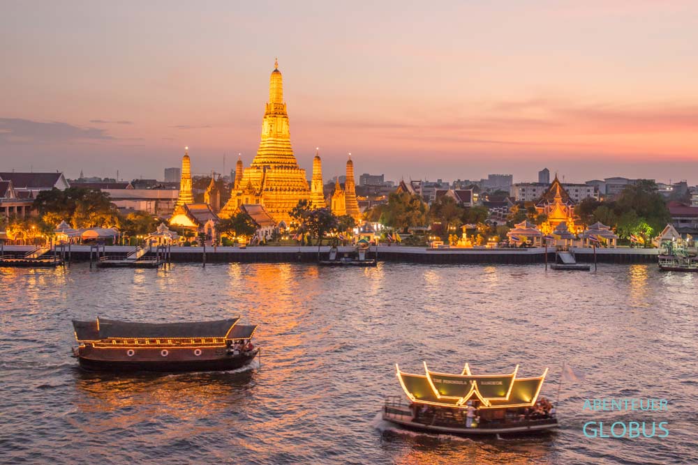 Wat Arun in Bangkok zur Abenddämmerung, beleuchtet am Chao-Phraya-Fluss, mit traditionellen Booten im Vordergrund.