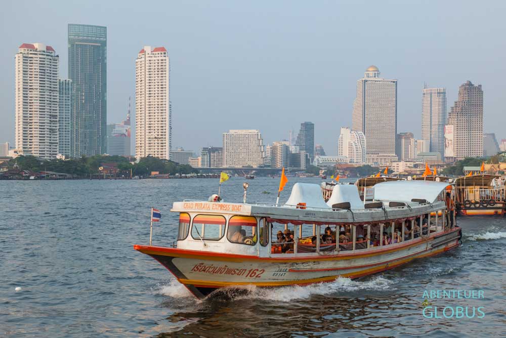 Ein Expressboot mit orangefarbenen Flaggen fährt über den Chao Phraya in Bangkok, dahinter die Skyline.