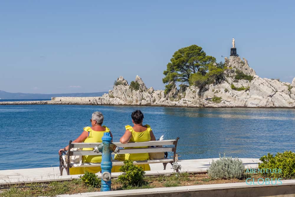 Felsen auf der Mole in Trpanj mit der Madonna-Statue Zvijezda Mora, im Vordergund zwei Frauen in gelben Kleidern auf einer Bank