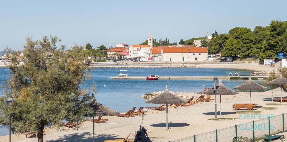 Strand Iza Banja mit Liegen und Sonnenschirmen in Sveti Filip i Jakov, dahinter die Altstadt mit dem Glockenturm der St.-Michael-Kirche.