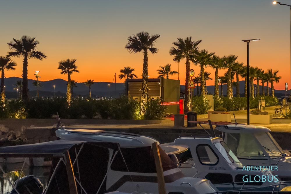 Hafen und Uferpromenade mit Palmen in Sveti Filip i Jakov. Im Hintergrund orangfarbener Abendhimmel über der Insel Pasman 