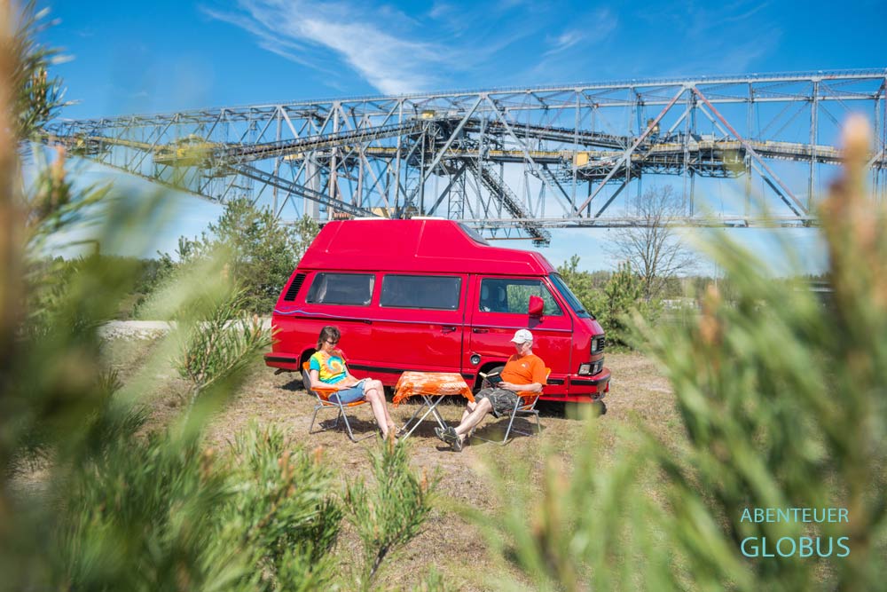 Rotes Wohnmobil VW T3, vor dem zwei Personen sitzen und lesen, im Hintergrund die Förderbrücke F60 in Lichterfeld-Schacksdorf