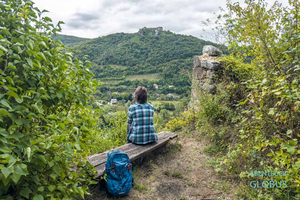 Urlauberin auf einer Bank an der Burgruine Havala. Im Hintergrund Festung Ostrovica auf grünem Berg in Kulen Vakuf im Una-Nationalpark