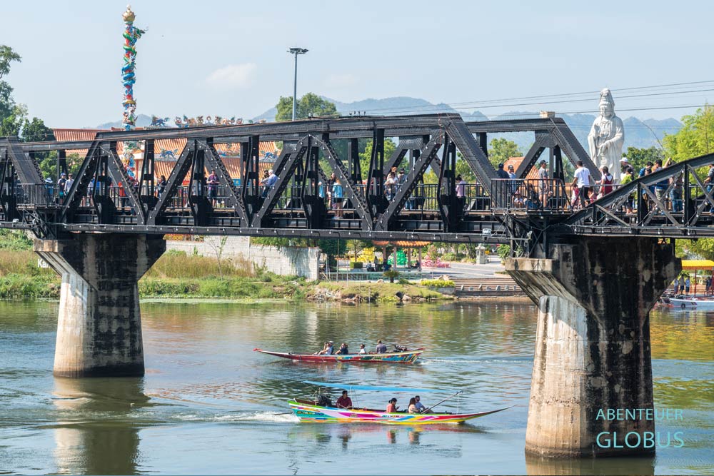 Ein Segment der Brücke am Kwai, auf dem Besucher spazieren, darunter zwei Boote mit Touristen auf dem Fluss in Kanchanaburi