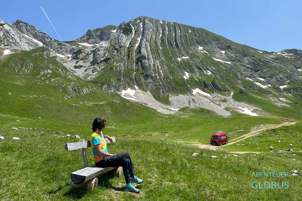 Besucherin auf einer Bank und roter Camper auf einem Parkplatz an der Panoramastraße Durmitor-Ring, im Hintergrund Berg Prutas im Frühjahr