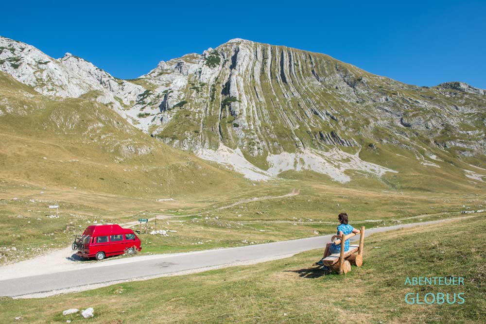 Besucherin auf einer Bank und roter Camper auf einem Parkplatz an der Panoramastraße Durmitor-Ring, im Hintergrund Berg Prutas im Herbst