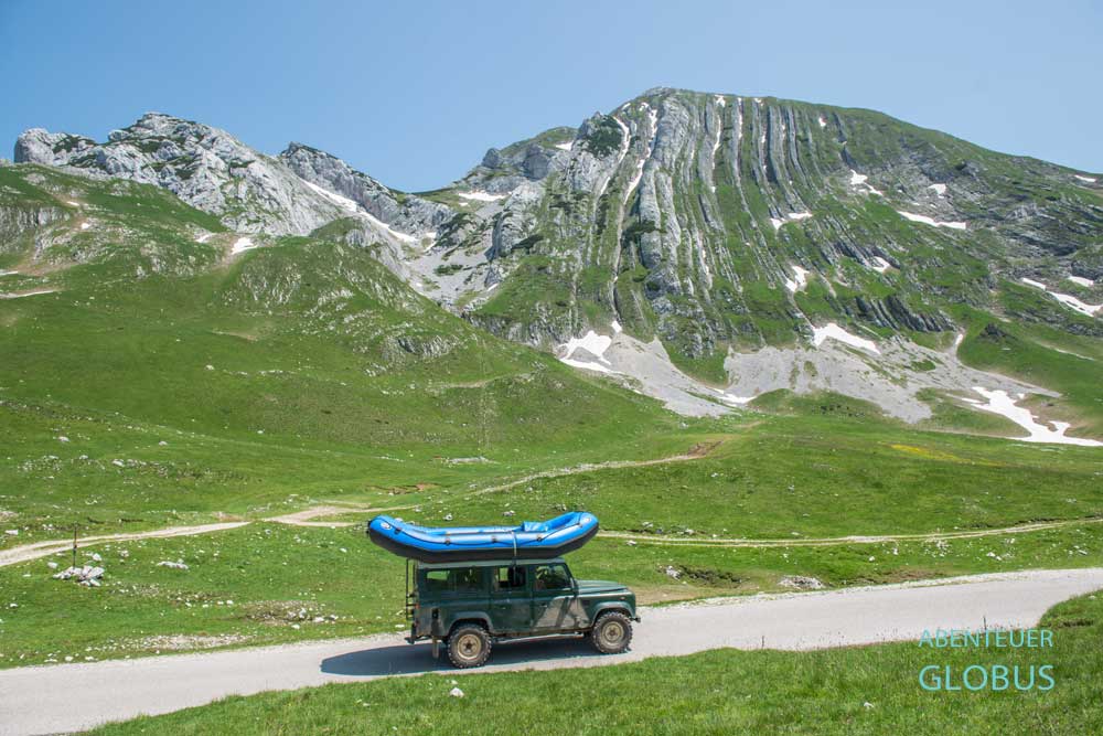 Jeep mit blauem Schlauchboot auf dem Dach vor der Bergkulisse des Prutas in Montenegro.