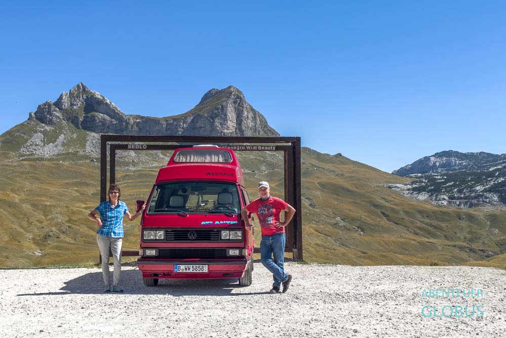 Zwei Touristen mit roten Van am Holzrahmen am Durmitor-Ring, im Hintergrund Bergmassiv Sedlena Greda