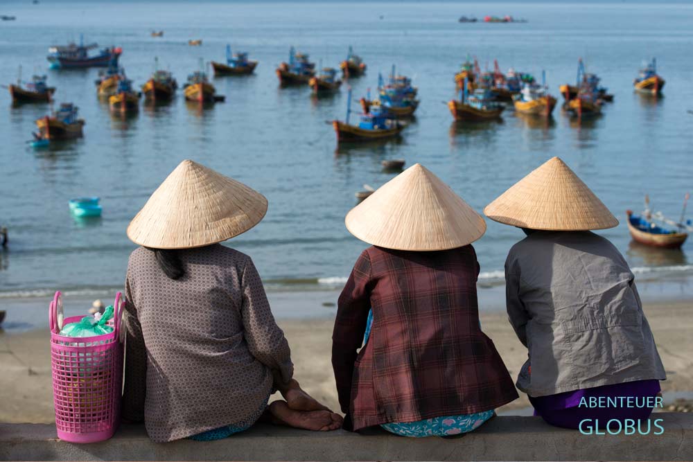 Drei Frauen mit Reishüten sitzen auf einer Mauer und blicken zu den Booten im Hafen von Mui Ne.