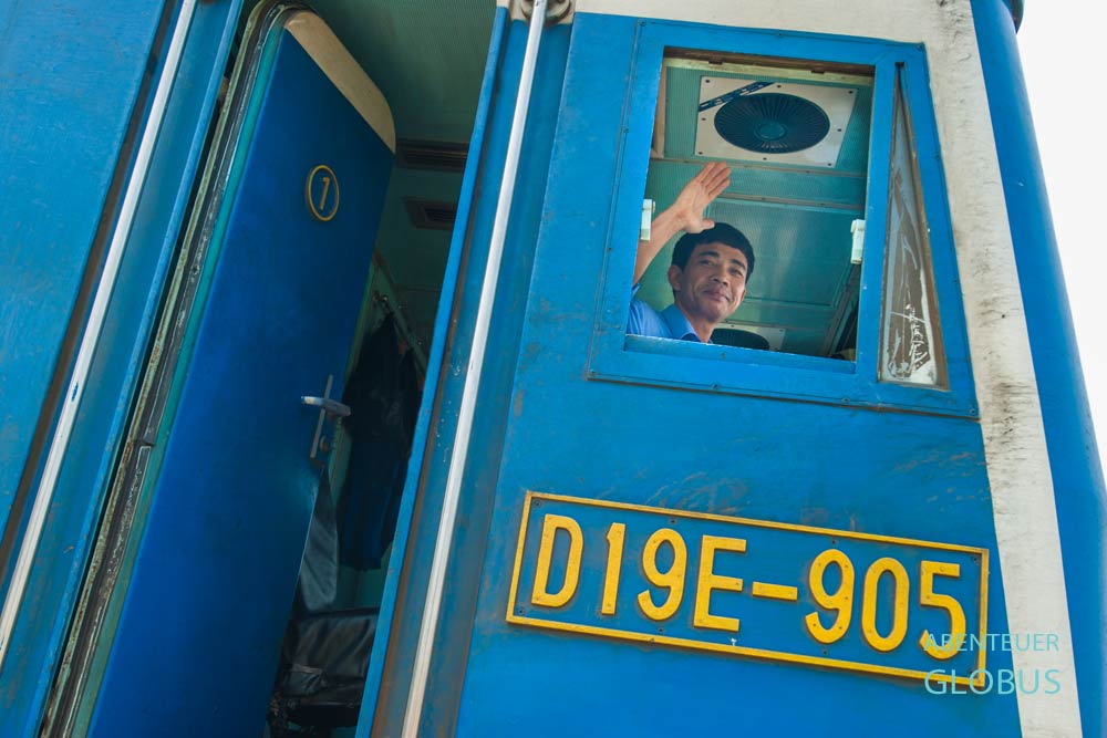 Lokführer winkt aus dem Fenster der Lok in die Kamera, unter ihm das Schild mit der Zugnummer D19E-905