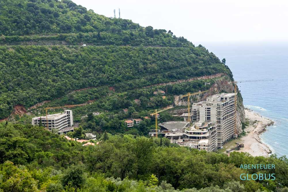 Bauruine des AS Hotels mit Baukränen am Strand Perazića Do unterhalb eines grünen Berges und der Adriamagistrale bei Petrovac na Moru, Montenegro