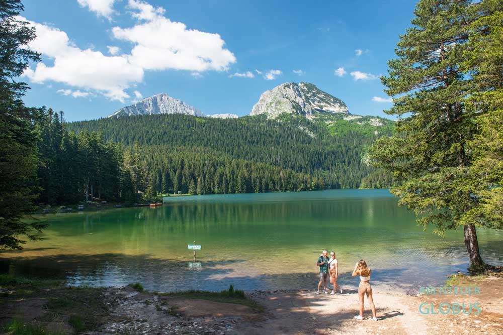 Drei Touristen am Ufer des Schwarzen Sees (Crno Jezero) und Blick zum Durmitormassiv mit Berg Bobotov Kuk bei Zabljak im Nationalpark Durmitor