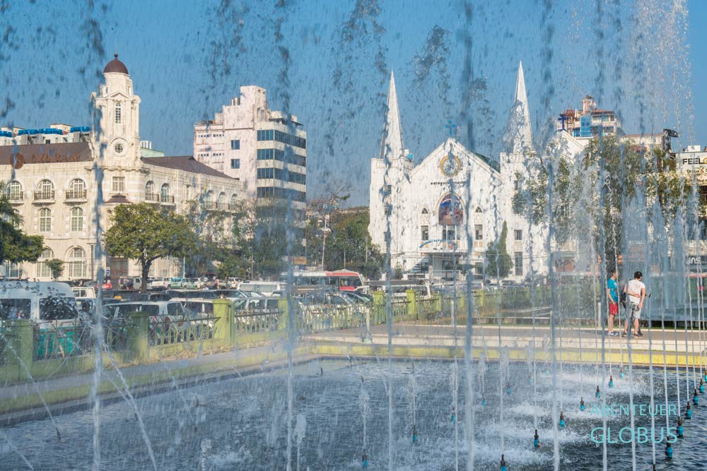 Blick auf einen Springbrunnen im Mahabandoola Garden, dahinter die AYA Bank und die Emmanuel Baptist Church in Yangon