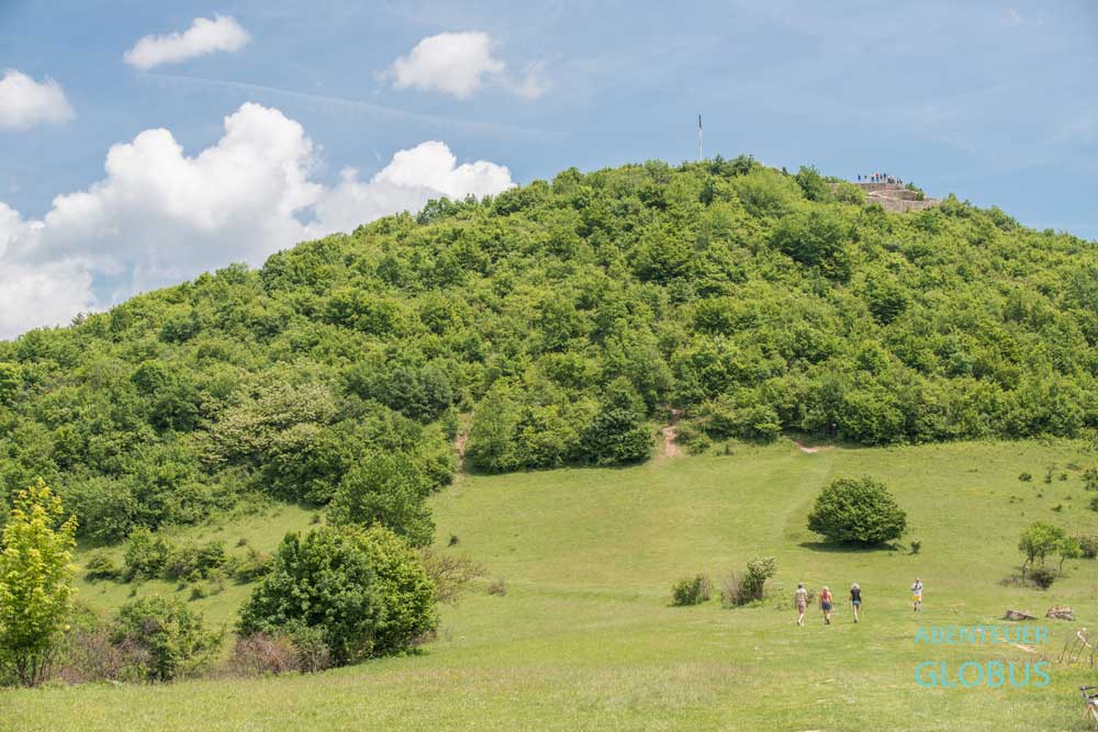 Am Fuße der Sonnenpyramide wandern Besucher über die Wiese. Auf dem Gipfel thront die Burgruine Stari Grad Visoko.