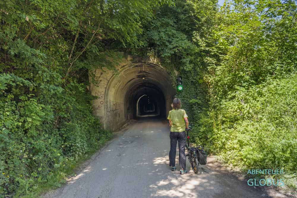 Radfahrerin am bewachsenen Eingang des ehemaligen Eisenbahntunnels auf der Seite am Kleinen Pliva-See in Jajce