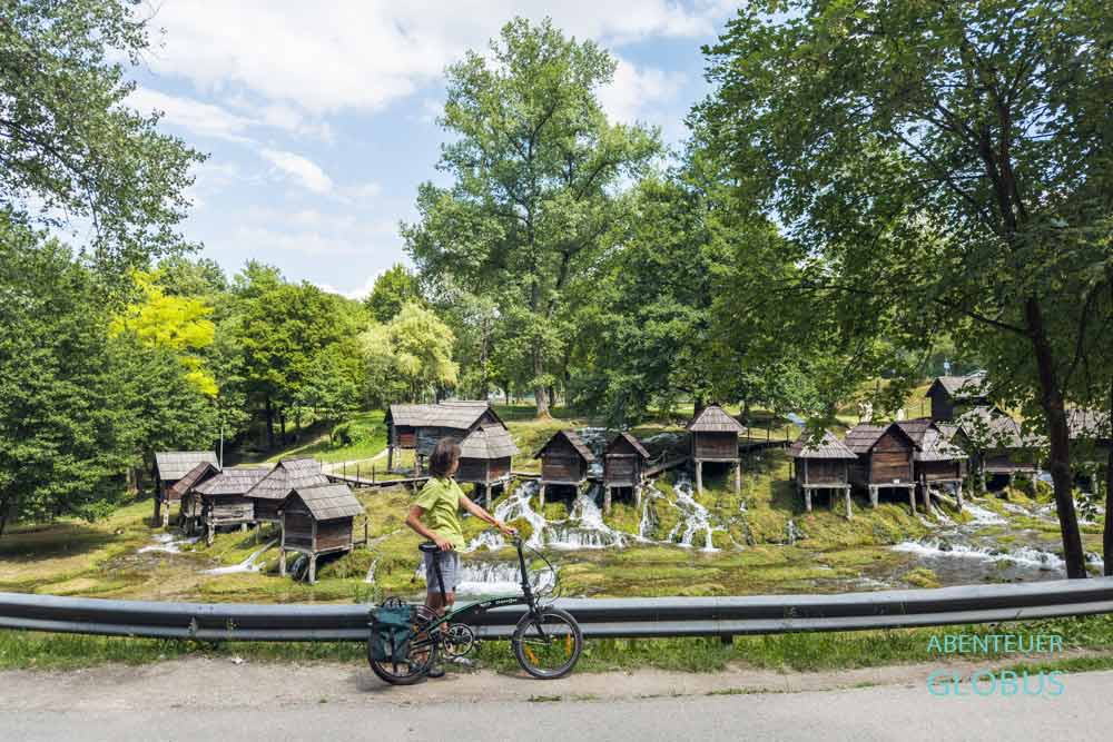 Radfahrerin am Straßenrand, dahinter die Wassermühlen Mlincici in Jajce