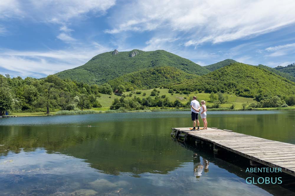 Zwei Personen auf dem Badesteg am See Dol, in dem sich die umliegenden grünen Berge spiegeln, in der Nähe von Jajce in Bosnien und Herzegowina