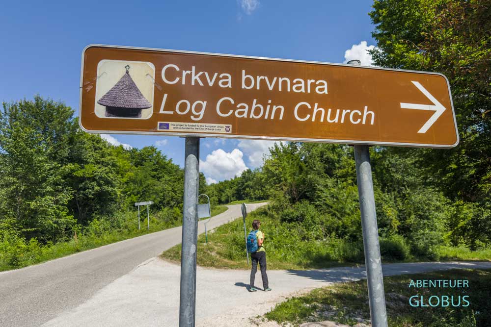 Hinweisschild zur Blockkirche St. Nikolaus, im Hintergrund eine Wanderin nahe Krupa na Vrbasu