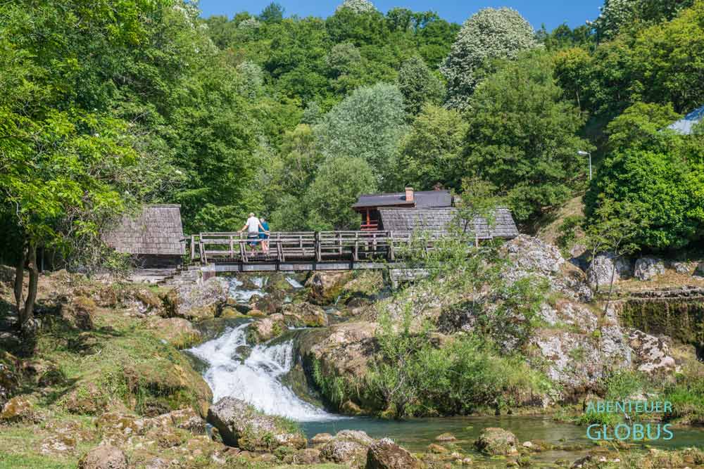 Zwei Touristen stehen auf der Holzbrücke an den Krupa Wasserfällen in Krupa na Vrbasu. Rechts und links sind alte Holzmühlen zu sehen.