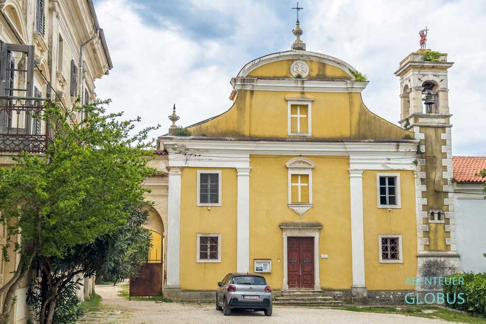 Gelbe, barocke Kirche des Heiligen Johannes des Täufers (Crkva Sv. Ivana Krstitelja) mit Glockenturm und parkenden Auto davor im Dorf Dajla bei Novigrad, Kroatien