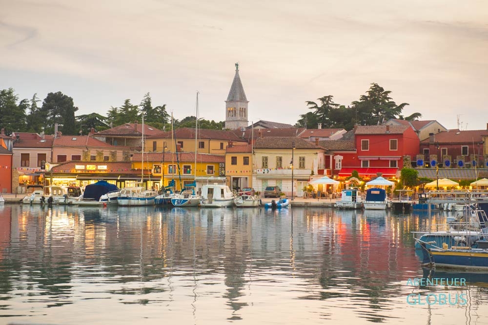 Beleuchteter Stadthafen Mandrač in Novigrad mit Promenade und Glockenturm der Pfarrkirche der Heiligen Pelagius und Maximus am Abend.