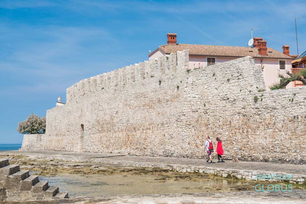 Stadtmauer, Promenade mit zwei Spaziergängern und Stadtstrand um die Altstadt von Novigrad auf der Halbinsel Istrien in Kroatien