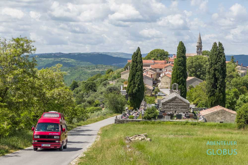 Roter Campervan auf einer Straße, Friedhof mit Kirche des heiligen Eliseus und das Dorf Draguc im Hintergrund