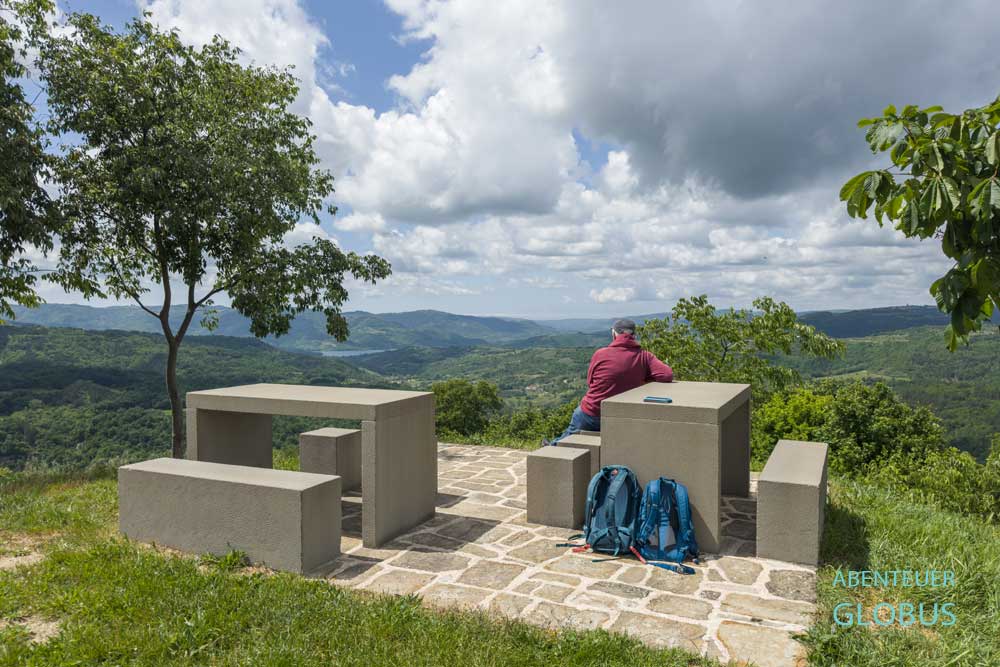 Zwei Betonsitzgruppen mit einem Touristen in Draguć, im Hintergrund Hügellandschaft und der Butoniga-See in Istrien.