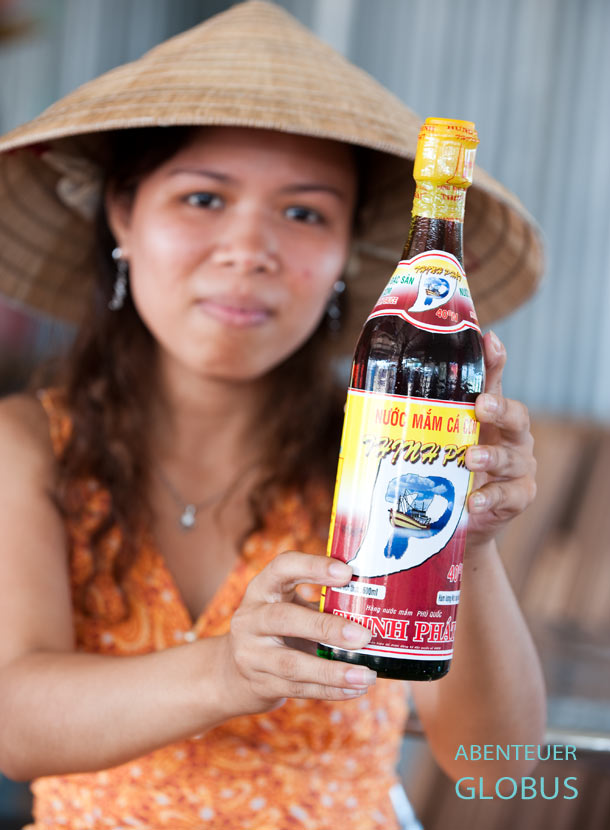 Eine vietnamesische Frau mit Strohhut hält eine volle Glasflasche Fischsoße in die Kamera.