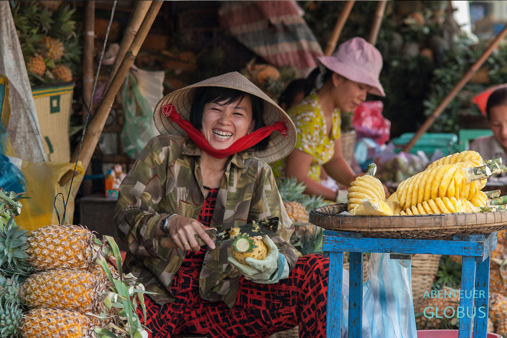 Vietnamesische Frau schält auf einem Markt eine Ananas und lacht dabei herzlich.
