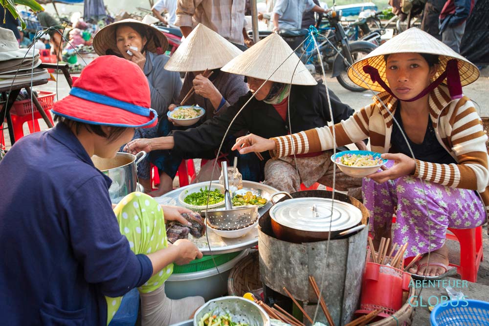 Vietnamesische Frauen sitzen auf kleinen Hockern an einem Streetfood-Stand und essen gemeinsam Nudelsuppe.