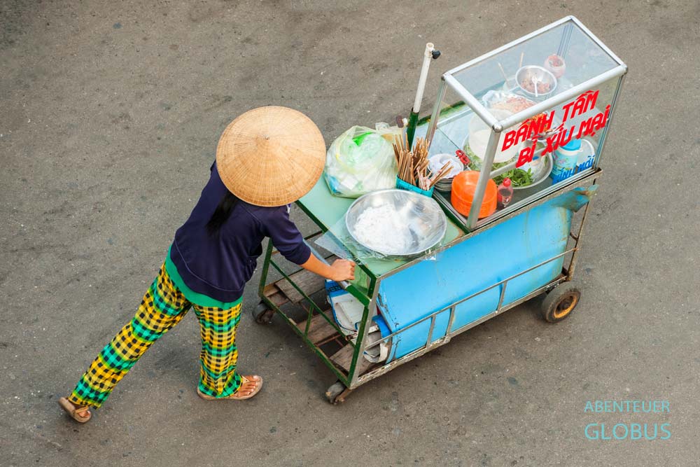 Vietnamesische Frau schiebt einen mobilen Streetfood-Stand auf der Straße, aufgenommen aus der Vogelperspektive.