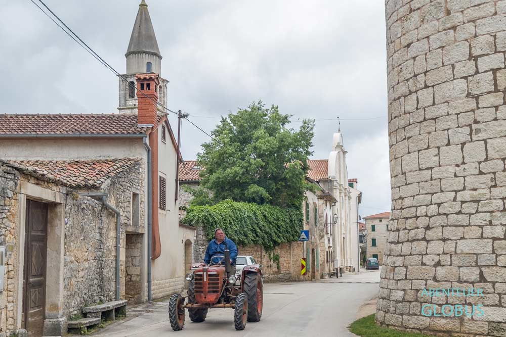 Roter Traktor mit Fahrer in einer Gasse, im Hintergrund die Pfarrkirche Maria Verkündigung