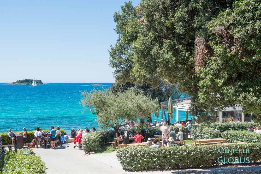 Urlauber im Restaurant mit Biergarten vom Campingplatz Porto Sole in der Brizine-Bucht, im Hintergrund Insel und Segelboot in der Adria in Vrsar