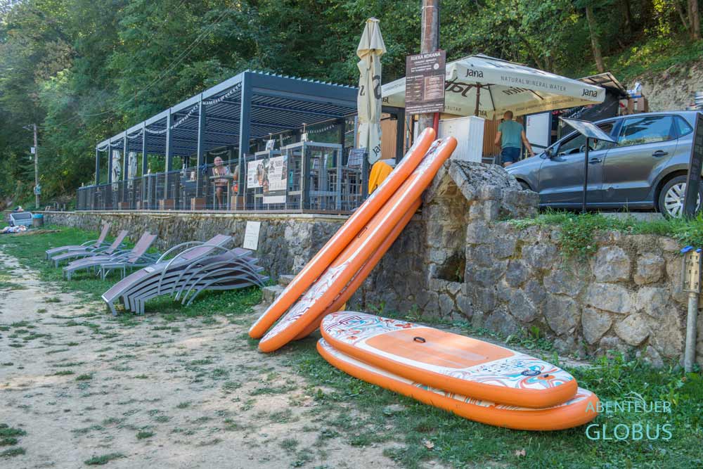 Vier orangefarbene SUP-Boards und graue Liegen, dahinter Beach Bar Taurus vom Korana Naturbad Slunj